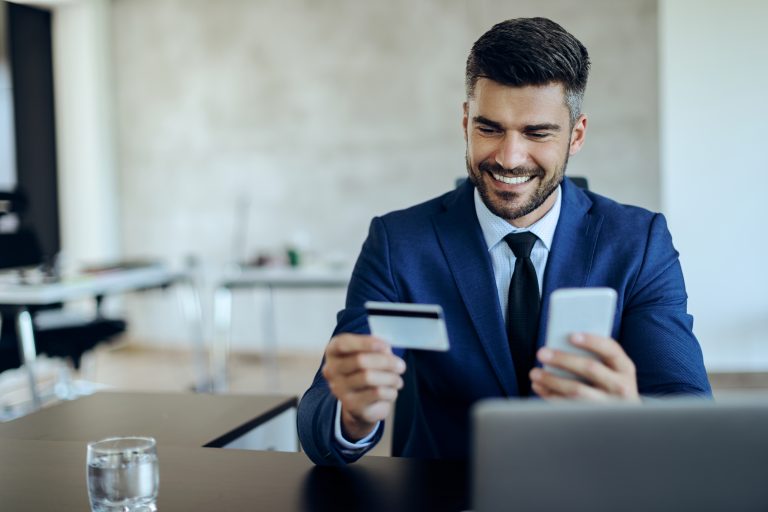 Smiling businessman in a blue suit sitting at a desk, holding a credit card in one hand and a smartphone in the other, managing finances online.
