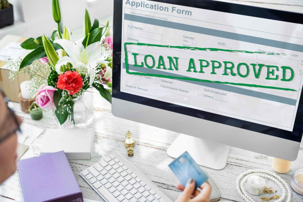 Approved commercial loan application form on computer screen with credit card in hand and flowers on desk