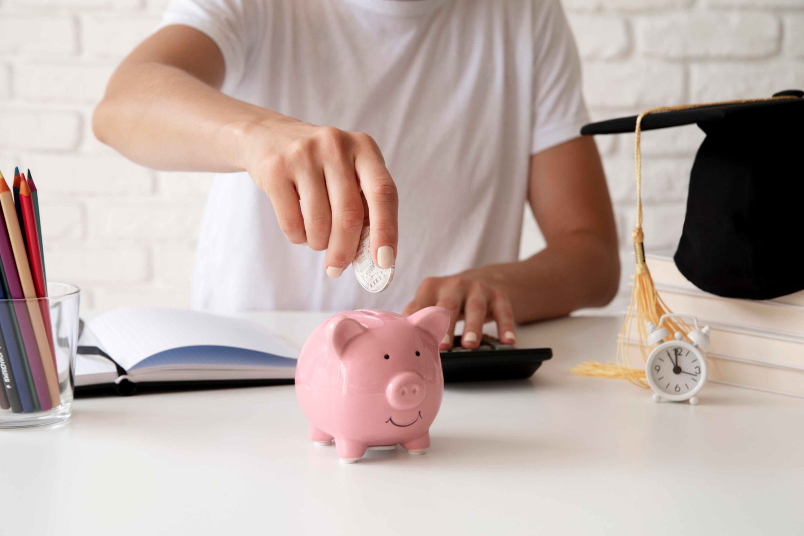 Person saving money by putting a coin into a pink piggy bank, with a graduation cap, alarm clock, calculator, and stationery on the desk — representing education savings, budgeting, or financial planning for students.