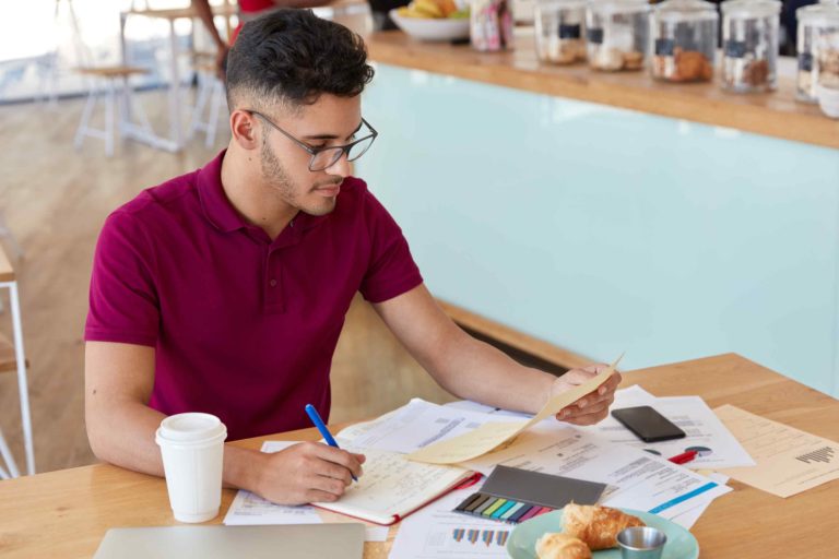 Young man in a maroon shirt reviewing financial documents at a table with charts, a notebook, coffee, and pastries, appearing focused on budgeting.