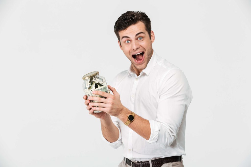 Excited young man in white shirt holding a jar full of cash, symbolizing smart spending and money-saving success.
